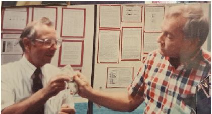 Two people facing each other; one hands the other a dollar bill. Papers of text are mounted to the wall behind them 